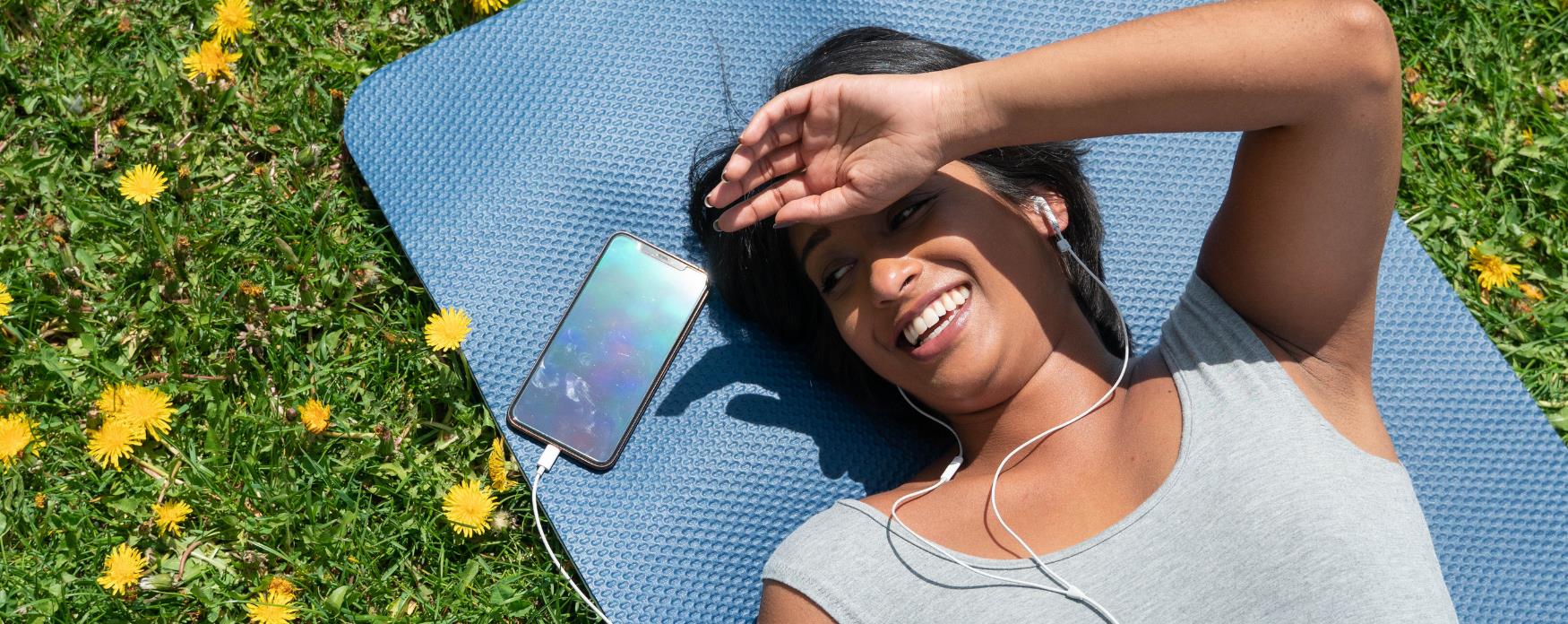 woman with headphones and phone lying on yoga mat in park