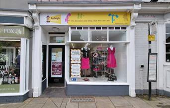 Outside of Farleigh Hospice charity shop in Maldon High Street with yellow sign and pink dresses in window