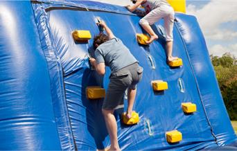 Two people climbing up a blue inflatable wall in a park